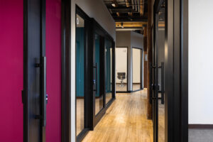 A hallway at The Village Salon Studios, showcasing several salon suites with large glass doors and colourful interiors. The hallway features polished wooden flooring and exposed industrial elements, creating a modern and inviting atmosphere. Suite 22 is visible at the end of the hallway with a styling chair inside.